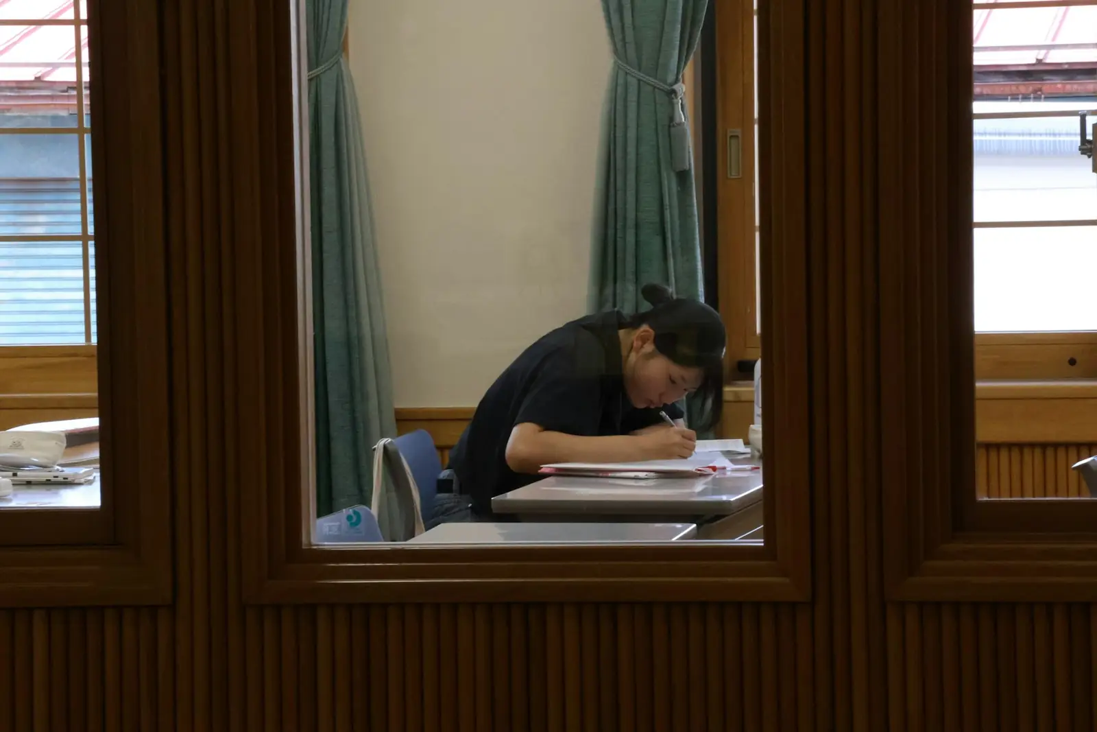 Ahşap pencere çerçevesinden görünen, masada ders çalışan öğrenci