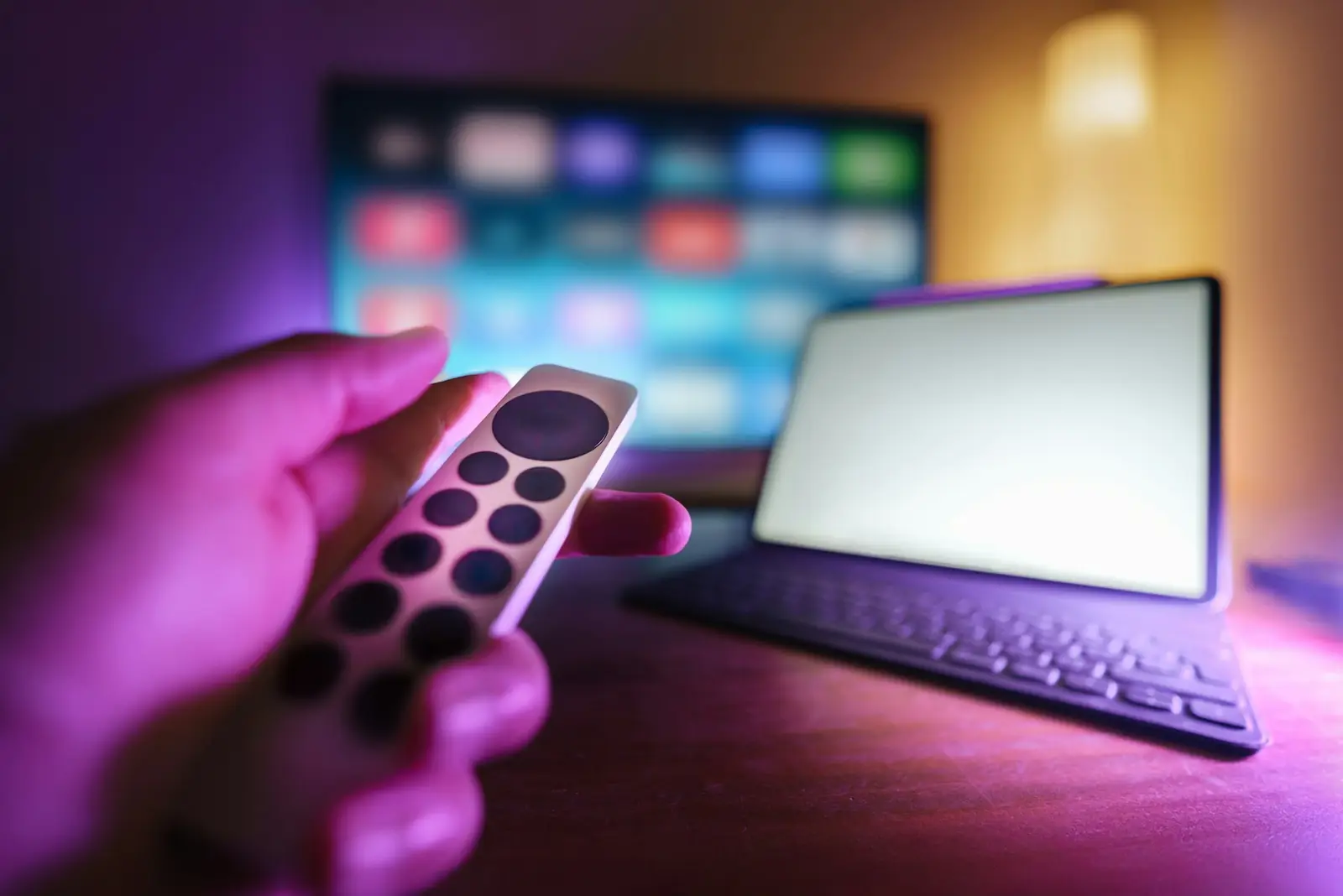 Mor ışıklı karanlık odada elinde TV kumandası tutan kişi, arkada bulanık akıllı TV ekranı ve tablet