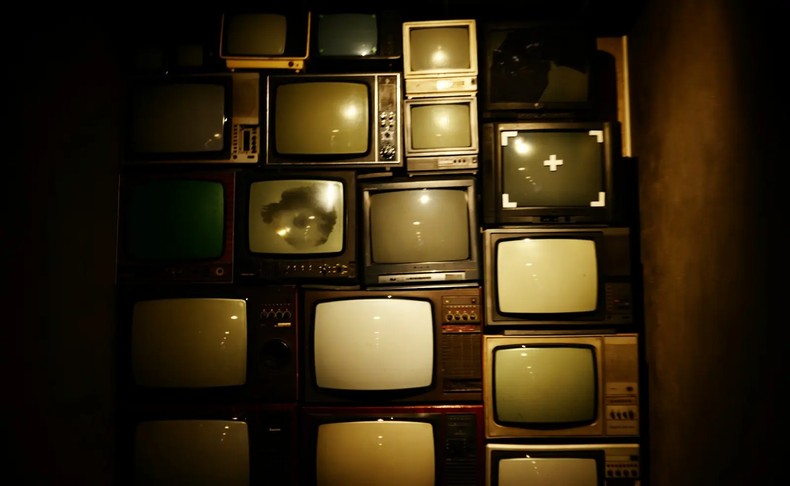 Loş ışıklı duvarda üst üste dizilmiş, ekranları yanan eski televizyonlar